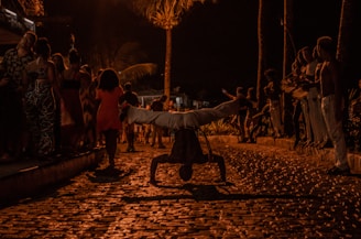 A night scene featuring a street performance with a person balancing on their hands. The background includes a group of people watching, some standing and others walking. The area is illuminated by warm street lights, casting shadows and creating a lively atmosphere.