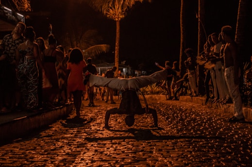 A night scene featuring a street performance with a person balancing on their hands. The background includes a group of people watching, some standing and others walking. The area is illuminated by warm street lights, casting shadows and creating a lively atmosphere.