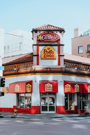 A fast-food restaurant located at a street corner features branding for both Carl's Jr. and El Pollo Loco. The building has a distinct red-tiled roof and prominent signage, with red and yellow color schemes. Red awnings adorn the windows and entrance, and the sidewalk outside is clean with some visible street elements like traffic lights and road signs.