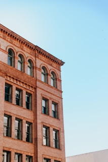 A multi-story brick building with large windows is depicted against a clear blue sky. The architectural style features arched windows on its upper floors and decorative brickwork along the edges.