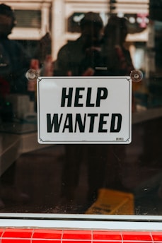 A 'Help Wanted' sign is prominently displayed on a glass window. The background shows a blurred view of the interior with some indistinct figures and objects, suggesting an indoor setting or a storefront.
