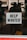 A 'Help Wanted' sign is prominently displayed on a glass window. The background shows a blurred view of the interior with some indistinct figures and objects, suggesting an indoor setting or a storefront.