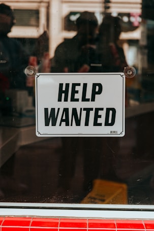 A 'Help Wanted' sign is prominently displayed on a glass window. The background shows a blurred view of the interior with some indistinct figures and objects, suggesting an indoor setting or a storefront.