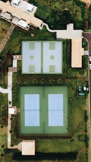 An aerial view showcases a residential area with two basketball courts and two tennis courts adjacent to each other. The courts are surrounded by grassy areas and several buildings with beige roofs. Pathways connect the various parts of the complex, and trees create a lush environment around the perimeter.