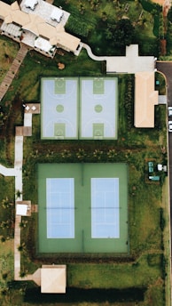 An aerial view showcases a residential area with two basketball courts and two tennis courts adjacent to each other. The courts are surrounded by grassy areas and several buildings with beige roofs. Pathways connect the various parts of the complex, and trees create a lush environment around the perimeter.