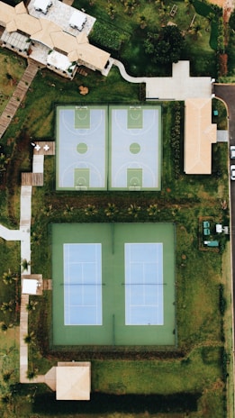 An aerial view showcases a residential area with two basketball courts and two tennis courts adjacent to each other. The courts are surrounded by grassy areas and several buildings with beige roofs. Pathways connect the various parts of the complex, and trees create a lush environment around the perimeter.