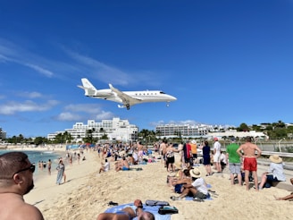 A large jet plane flies low over a crowded beach, with numerous people watching and relaxing on the sand. The beachgoers are dressed in swimwear, enjoying a sunny day. In the background, a resort with palm trees and multi-story buildings is visible.