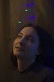 A person gazes upward with a contemplative expression, illuminated by soft, muted lighting. The background features subtle, colorful lights that add a dreamy atmosphere.