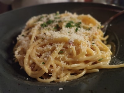 A plate of creamy pasta topped with grated cheese and garnished with chopped herbs, placed in a dark-colored dish. The pasta is well-coated with a cheesy sauce, and the texture appears rich and smooth.