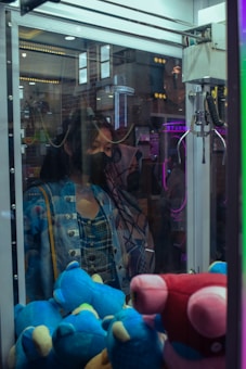 A person wearing a mask stands in front of a claw machine filled with colorful plush toys. The reflections in the glass show a dimly lit entertainment area with neon lights.