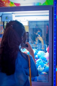 A person with long hair is standing in front of a claw machine filled with plush toys, taking a photo with a smartphone. The reflection of the individual can be seen in the glass of the machine. The plush toys inside are primarily blue, and there's bright, colorful decor on the top of the machine.