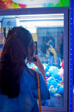 A person with long hair is standing in front of a claw machine filled with plush toys, taking a photo with a smartphone. The reflection of the individual can be seen in the glass of the machine. The plush toys inside are primarily blue, and there's bright, colorful decor on the top of the machine.