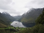 A serene landscape featuring a fjord surrounded by lush green valleys and towering mountains partially covered in snow. A cruise ship is visible in the fjord, and small villages are nestled below the mountains. The sky is overcast, adding a moody and peaceful atmosphere.