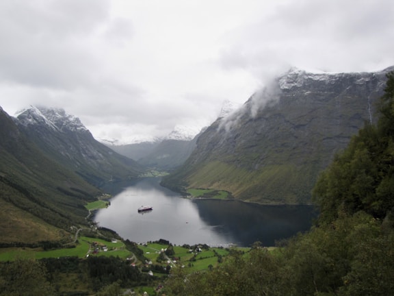 A serene landscape featuring a fjord surrounded by lush green valleys and towering mountains partially covered in snow. A cruise ship is visible in the fjord, and small villages are nestled below the mountains. The sky is overcast, adding a moody and peaceful atmosphere.