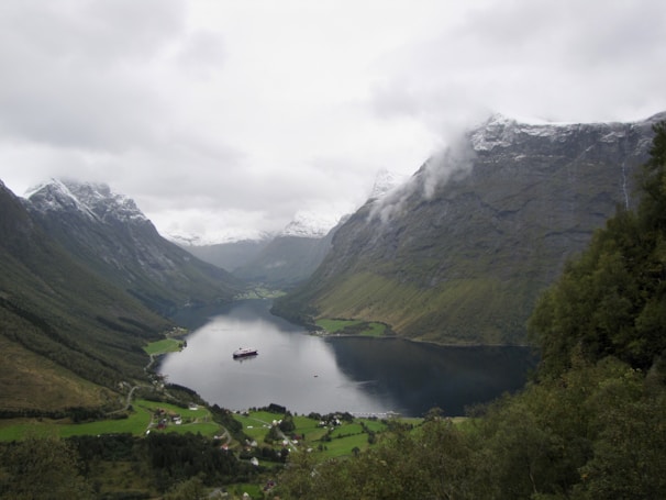 A serene landscape featuring a fjord surrounded by lush green valleys and towering mountains partially covered in snow. A cruise ship is visible in the fjord, and small villages are nestled below the mountains. The sky is overcast, adding a moody and peaceful atmosphere.