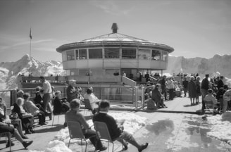 A group of people gathered on an outdoor terrace in front of a circular building with large windows. Snow-covered mountains surround the area, and several individuals are sitting on chairs, conversing, or walking around. The atmosphere is lively and social.