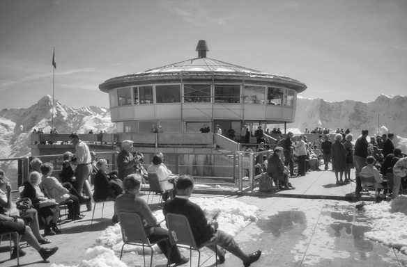 A group of people gathered on an outdoor terrace in front of a circular building with large windows. Snow-covered mountains surround the area, and several individuals are sitting on chairs, conversing, or walking around. The atmosphere is lively and social.