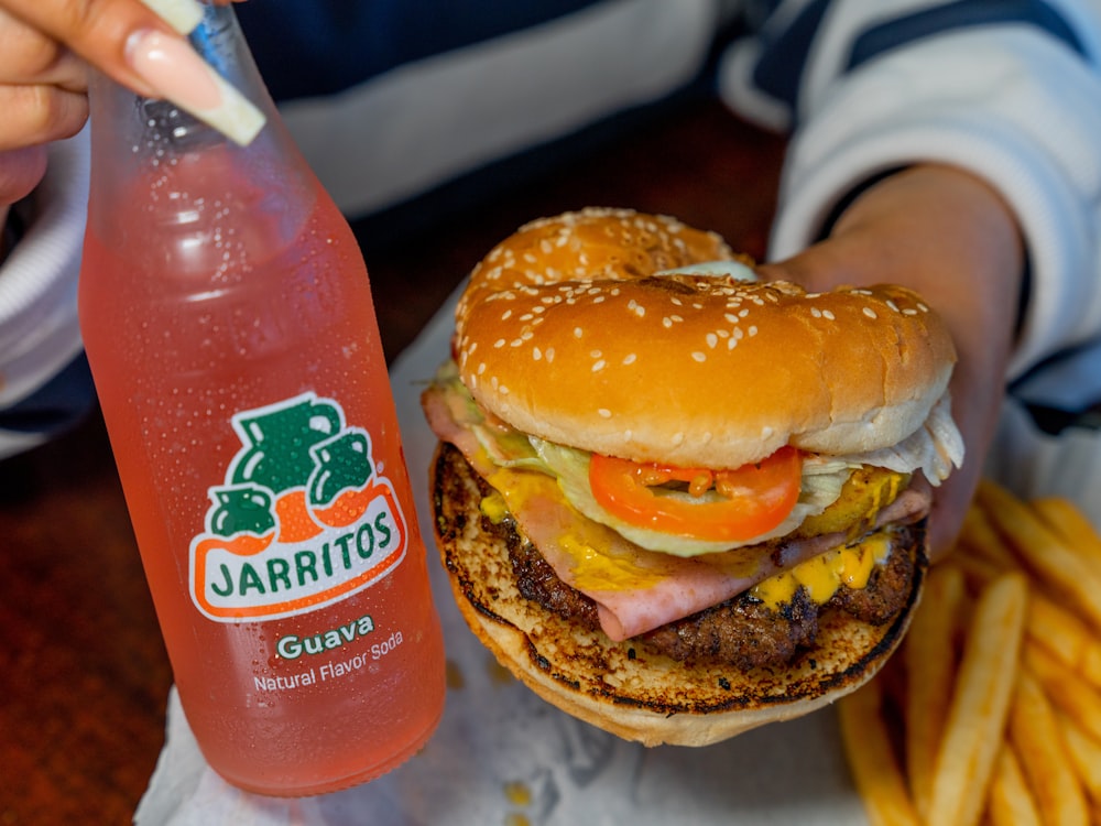 uma pessoa segurando um hambúrguer grande ao lado de uma garrafa de suco