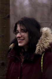 A person with glasses and long dark hair is smiling. They are wearing a maroon jacket with a fur-lined hood. The background is a blurred, natural setting with hints of tree trunks.