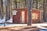 A small, rustic wooden cabin is situated in a forested area during winter. The cabin is adorned with various vintage license plates on its exterior. Snow covers the ground surrounding the cabin, while tall pine trees envelop the structure, casting shadows on the snow.