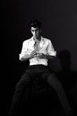 A black and white portrait featuring a person seated on a chair, wearing a partially unbuttoned white shirt and dark pants. The individual has a serious expression, with slightly tousled hair and a focused gaze. The lighting creates a dramatic contrast and casts a distinct shadow on the backdrop.
