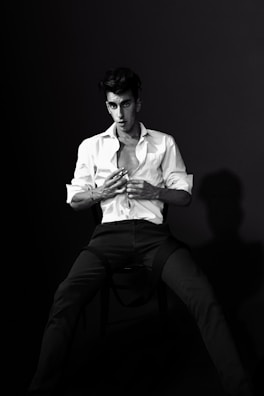 A black and white portrait featuring a person seated on a chair, wearing a partially unbuttoned white shirt and dark pants. The individual has a serious expression, with slightly tousled hair and a focused gaze. The lighting creates a dramatic contrast and casts a distinct shadow on the backdrop.