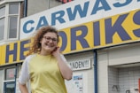 A person with curly hair and glasses is smiling in front of a car wash sign. The sign is large with blue and yellow colors. The building is made of brick, and there are small posters and signs nearby.