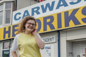 A person with curly hair and glasses is smiling in front of a car wash sign. The sign is large with blue and yellow colors. The building is made of brick, and there are small posters and signs nearby.