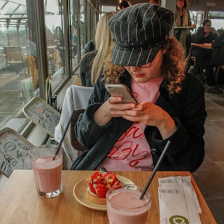A person wearing a hat is focused on their smartphone while seated at a wooden table in a cafe. The table has two glasses of pink smoothie and a plate with a strawberry-topped dessert. The cafe has large windows, allowing natural light to illuminate the setting.