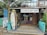 A rustic surf school is situated in a tropical setting, featuring surfboards displayed outside. The building is made of wood with a thatched roof, and there are signs advertising surfing lessons, accessories, and board repairs. Palms and tropical plants surround the school, contributing to a laid-back, beachy atmosphere.