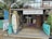 A rustic surf school is situated in a tropical setting, featuring surfboards displayed outside. The building is made of wood with a thatched roof, and there are signs advertising surfing lessons, accessories, and board repairs. Palms and tropical plants surround the school, contributing to a laid-back, beachy atmosphere.