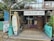 A rustic surf school is situated in a tropical setting, featuring surfboards displayed outside. The building is made of wood with a thatched roof, and there are signs advertising surfing lessons, accessories, and board repairs. Palms and tropical plants surround the school, contributing to a laid-back, beachy atmosphere.