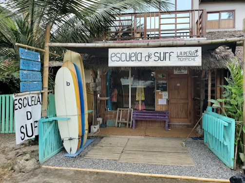 A rustic surf school is situated in a tropical setting, featuring surfboards displayed outside. The building is made of wood with a thatched roof, and there are signs advertising surfing lessons, accessories, and board repairs. Palms and tropical plants surround the school, contributing to a laid-back, beachy atmosphere.