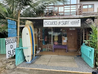 A rustic surf school is situated in a tropical setting, featuring surfboards displayed outside. The building is made of wood with a thatched roof, and there are signs advertising surfing lessons, accessories, and board repairs. Palms and tropical plants surround the school, contributing to a laid-back, beachy atmosphere.