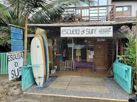 A rustic surf school is situated in a tropical setting, featuring surfboards displayed outside. The building is made of wood with a thatched roof, and there are signs advertising surfing lessons, accessories, and board repairs. Palms and tropical plants surround the school, contributing to a laid-back, beachy atmosphere.
