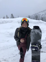 A welcoming ski instructor holding a snowboard, smiling against a snowy mountain backdrop.