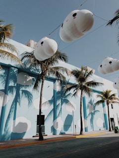Large, cute cloud-shaped balloons are suspended in the air against a backdrop of palm trees. The building in the scene is painted sky blue with white cloud-like patterns, creating a whimsical and playful atmosphere. The shadows of the palm trees are cast onto the building, adding depth to the composition.