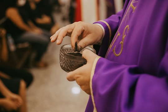 A person wearing a purple robe adorned with gold embroidery is holding a small, ornate glass container with black substance inside. The scene suggests a ceremonial or religious context, possibly involving a ritual.