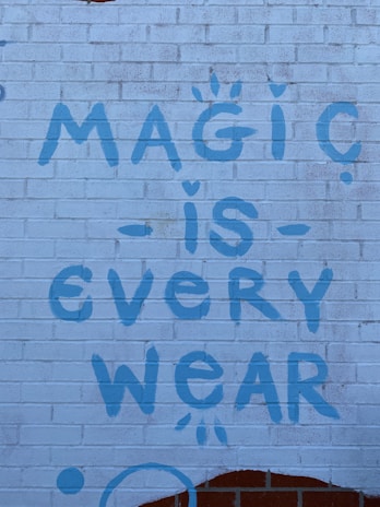 A brick wall with a graffiti-style message painted in blue, reading 'Magic is Everywhere' with playful symbols like dots and small stars accenting the letters.