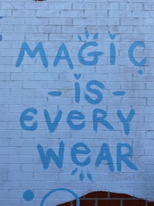 A brick wall with a graffiti-style message painted in blue, reading 'Magic is Everywhere' with playful symbols like dots and small stars accenting the letters.
