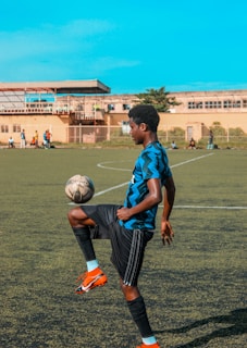 A young man is skillfully balancing a soccer ball on his knee on an outdoor soccer field. He is wearing a blue and black striped jersey, black shorts, and bright orange soccer cleats. The field is bordered by a building in the background where other players can be seen standing. The sky is clear and blue, suggesting a sunny day.