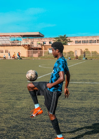 A young man is skillfully balancing a soccer ball on his knee on an outdoor soccer field. He is wearing a blue and black striped jersey, black shorts, and bright orange soccer cleats. The field is bordered by a building in the background where other players can be seen standing. The sky is clear and blue, suggesting a sunny day.