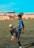 A young man is skillfully balancing a soccer ball on his knee on an outdoor soccer field. He is wearing a blue and black striped jersey, black shorts, and bright orange soccer cleats. The field is bordered by a building in the background where other players can be seen standing. The sky is clear and blue, suggesting a sunny day.