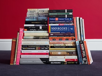 A collection of books is neatly stacked against a bold red wall. The books are arranged in two adjacent piles of varying heights, with a mix of colors and designs on the covers. Some books are arranged horizontally, while others are vertically aligned. The titles and various books cover a range of topics and genres.