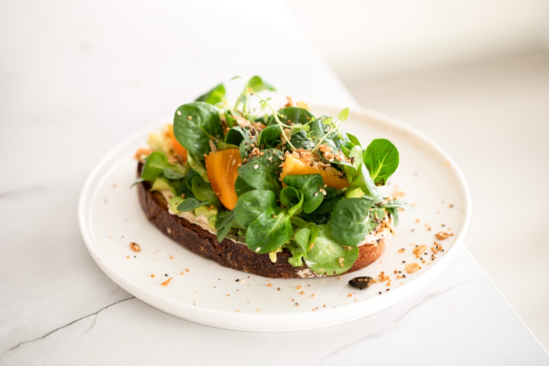 a white plate topped with a piece of bread covered in veggies, Vegan Cream Cheese & Avocado Toast with Lamb
