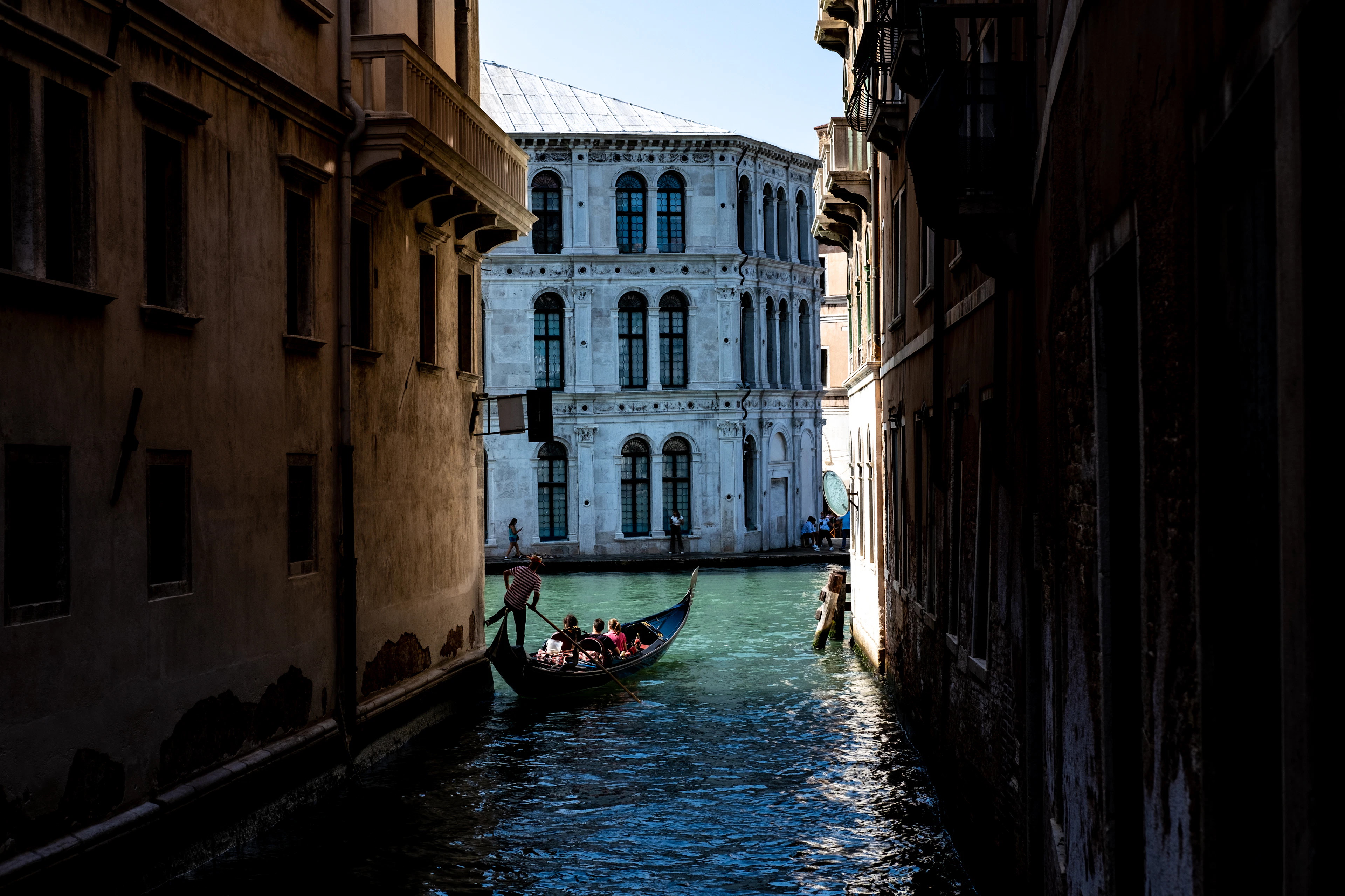 A gondolier steering a gondola through a shadowed back canal in Venice, a sunlit stone palazzo facade at the far end of the waterway