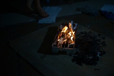 A small controlled fire burns inside a container filled with crumpled paper. The fire emits a warm glow, contrasting with the dark surroundings. Pieces of burnt paper or ashes are scattered nearby. Part of a person's foot wearing sandals is visible in the background along with scattered papers on the floor.
