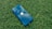 A blue smartphone with a visible Apple logo lies on a textured green grass surface. The phone's surface reflects a cloudy sky.