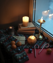 View of snow falling outside a window with candles and books on the sill, casting a warm glow inside.