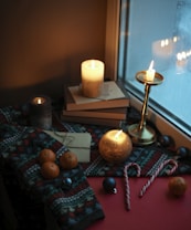 A cozy winter setting near a window, featuring lit candles on top of stacked books. A patterned knit blanket covers part of the scene, with several small oranges placed on it. Nearby are letters wrapped with string and candy canes. The warm glow from the candles is reflected in the window, enhancing the intimate ambiance.